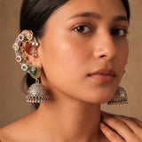 Indian model with loose dark waves wearing dual tone Navratna floral ear cuff jhumka on left ear, warm beige editorial portrait showing full crescent cuff from helix to lobe, green meenakari heart and oversized filigree jhumka