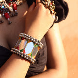 Close-up of wrist on wooden railing wearing large mirror tile ghungroo cuff bracelet outdoors with green leaves background, showing all mirror panels, thread zigzag detail and ghungroo ball borders clearly