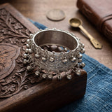 Silver wide filigree bangle with ghungroo drops on green marble surface with blue lapis lazuli, red ruby gemstones and ancient silver coin beside block-printed indigo textile, museum-style heritage flat lay