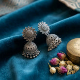Pair of oxidized silver sunflower jhumka earrings on teal velvet with dried rose buds and brass box, aesthetic flat lay