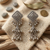 Oxidized silver diamond square chandelier jhumka earrings on rust red fabric with star anise, pearl string and teal brocade flat lay