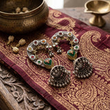 Dual tone Navratna floral ear cuff jhumka pair laid flat on deep maroon Banarasi zari brocade fabric with carved wooden block, antique brass singing bowl and small brass diya, heritage temple-style jewellery flat lay