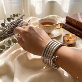 Silver oxidized 5-coil snake wrap bracelet laid flat in circle on ivory satin fabric with eucalyptus leaves and lavender dish, lifestyle flat lay showing full coil structure and ghungroo charm