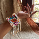 Close-up of hand wearing Kutchi embroidery cowrie cuff bracelet against terracotta wall, showing mirror shisha tiles, multicolour thread diamond motifs and pearl bead cowrie fringe drops in detail