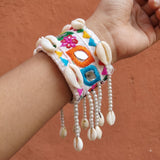 Hand raised near linen drape window wearing Kutchi embroidery mirror work cowrie cuff bracelet, natural light showing full cuff width and swaying pearl bead fringe with cowrie drops