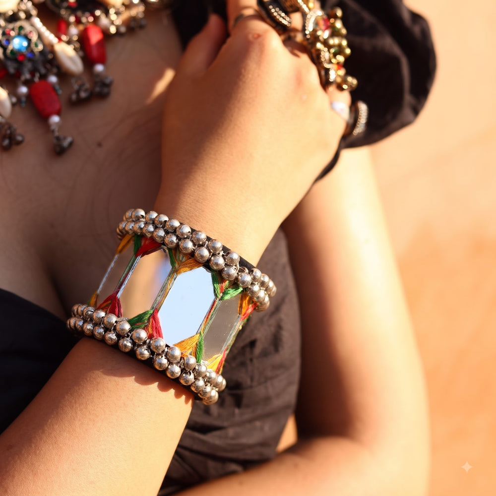 Close-up of wrist on wooden railing wearing large mirror tile ghungroo cuff bracelet outdoors with green leaves background, showing all mirror panels, thread zigzag detail and ghungroo ball borders clearly