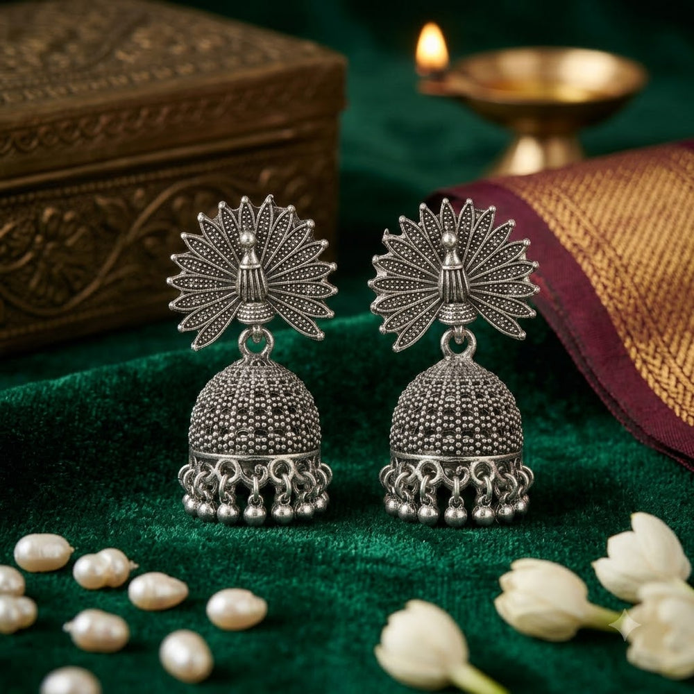 Pair of oxidized silver peacock jhumka earrings on green velvet with white jasmine flowers, freshwater pearls and brass diya, Diwali festive flat lay
