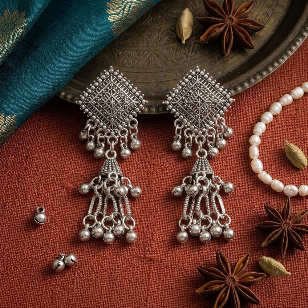 Pair of oxidized silver diamond square chandelier jhumka earrings on cream woven fabric with river stones and dried leaf, aesthetic flat lay