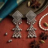 Pair of oxidized silver diamond square chandelier jhumka earrings on cream woven fabric with river stones and dried leaf, aesthetic flat lay