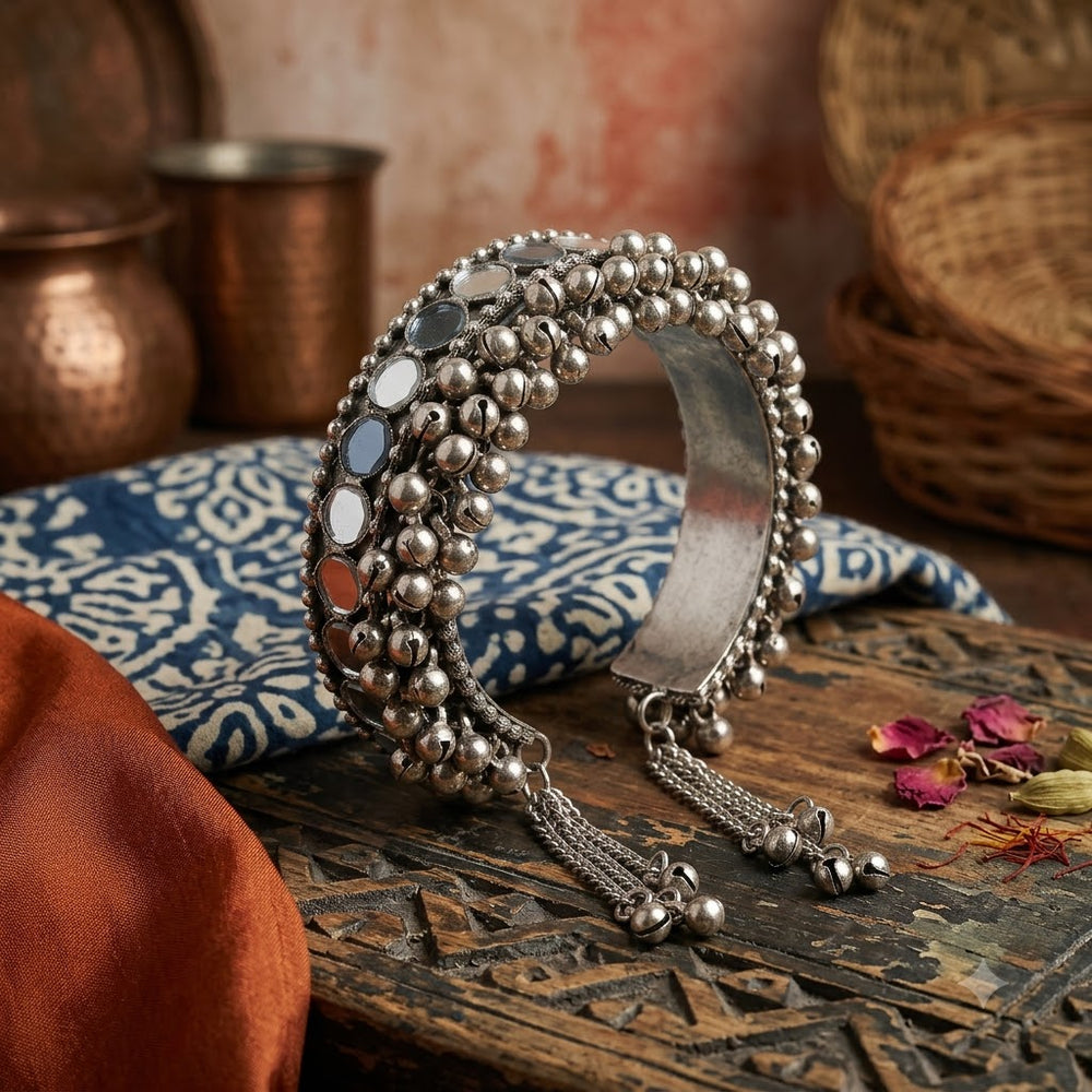 Oxidized silver ghungroo mirror cuff bangle resting on carved dark wood tray with indigo block-print fabric, orange silk, copper glass and rose petals, heritage aesthetic flat lay