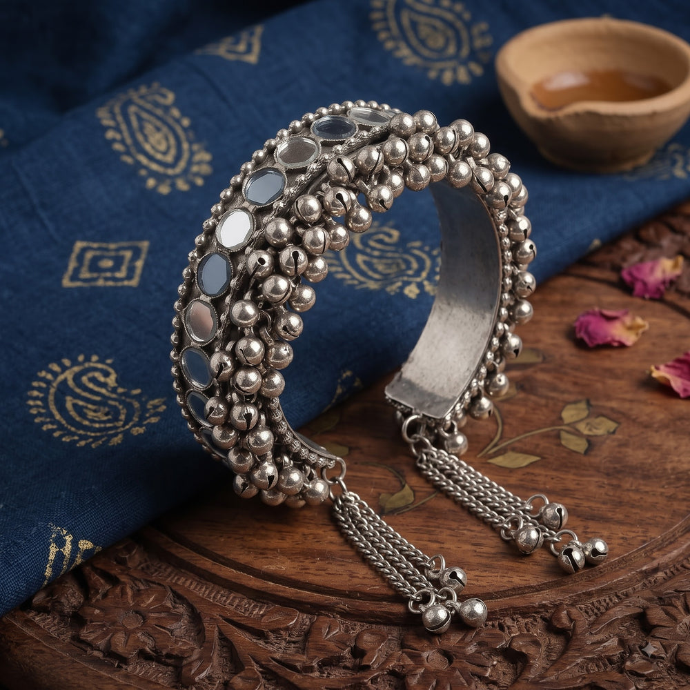 Oxidized silver ghungroo mirror cuff bangle on blue zari paisley block-print fabric with wooden diya bowl and dried rose petals, detail flat lay showing ghungroo texture, mirror tiles and chain tassel