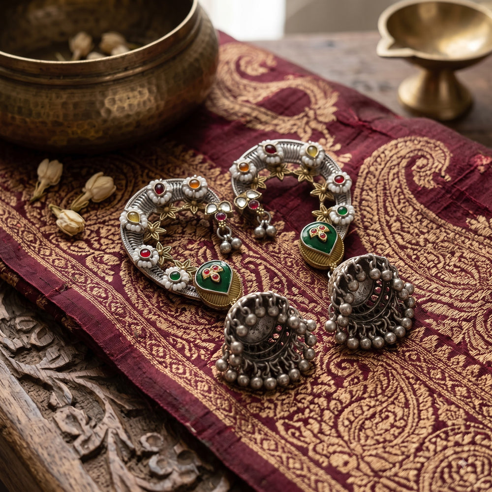 Dual tone Navratna floral ear cuff jhumka pair laid flat on deep maroon Banarasi zari brocade fabric with carved wooden block, antique brass singing bowl and small brass diya, heritage temple-style jewellery flat lay