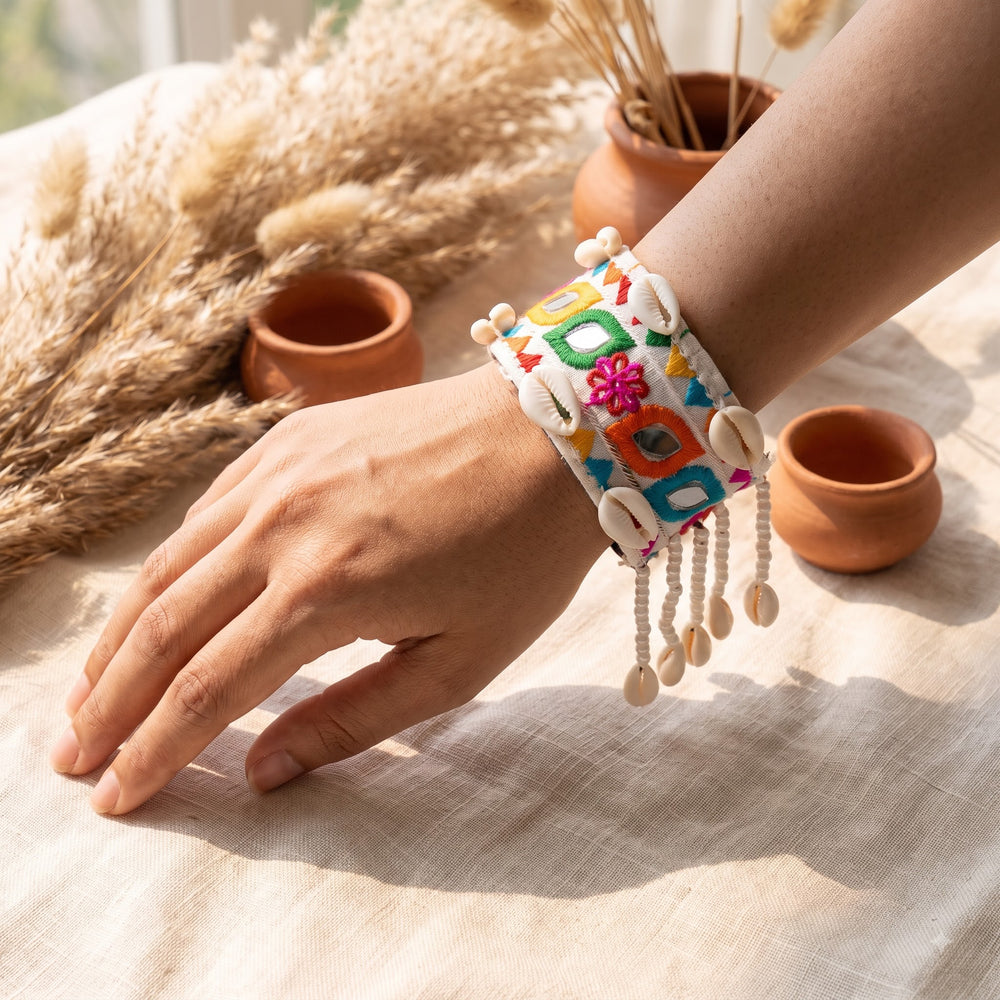 Kutchi embroidery mirror work cowrie shell cuff bracelet on wrist with pampas grass, terracotta pots and linen fabric in warm boho flat lay, showing full pearl bead and cowrie fringe