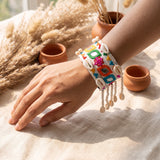 Kutchi embroidery mirror work cowrie shell cuff bracelet on wrist with pampas grass, terracotta pots and linen fabric in warm boho flat lay, showing full pearl bead and cowrie fringe