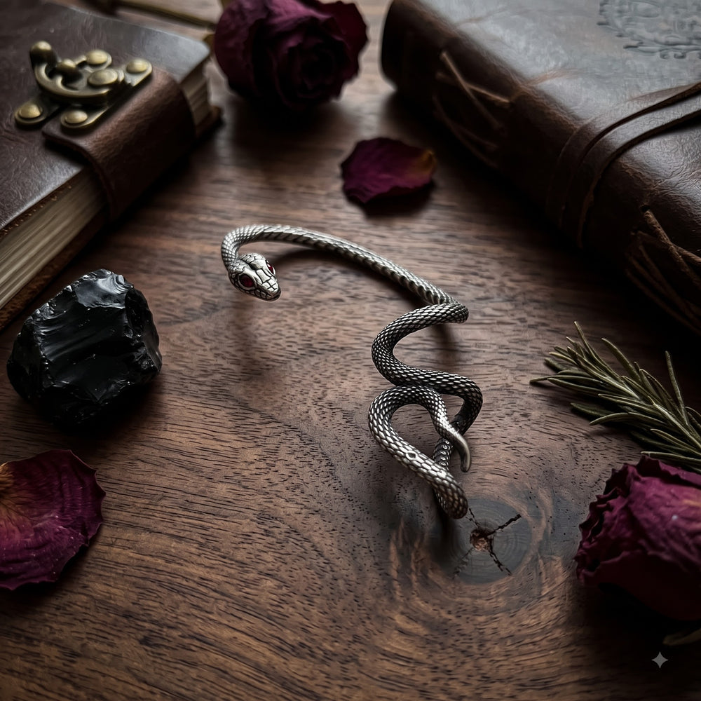 Oxidized silver snake ear cuff with red gemstone eyes on dark grey ear mannequin, blue damask fabric background with vintage jewellery box, garnet stones, magnifying glass and dried dark rose, gothic dark academia flat lay