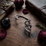 Oxidized silver snake ear cuff with red gemstone eyes on dark grey ear mannequin, blue damask fabric background with vintage jewellery box, garnet stones, magnifying glass and dried dark rose, gothic dark academia flat lay