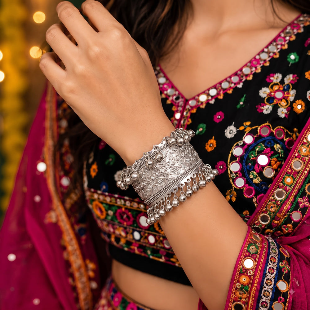 Close-up of wrist wearing silver wide filigree bangle with ghungroo drops in natural outdoor light, showing raised filigree oval motifs and crown edge with chiming silver ghungroo bells