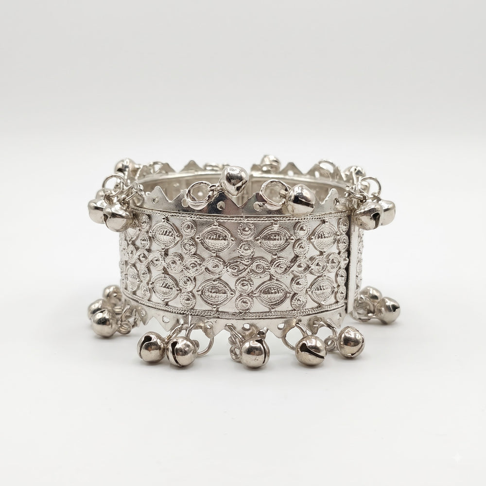 Silver wide filigree bangle with crown zigzag top edge and ghungroo drops on plain white background, front view showing raised repoussé oval spiral motifs and ghungroo bells on top and bottom rims