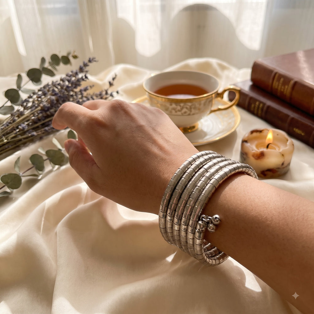 Silver oxidized 5-coil snake wrap bracelet laid flat in circle on ivory satin fabric with eucalyptus leaves and lavender dish, lifestyle flat lay showing full coil structure and ghungroo charm