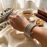 Silver oxidized 5-coil snake wrap bracelet laid flat in circle on ivory satin fabric with eucalyptus leaves and lavender dish, lifestyle flat lay showing full coil structure and ghungroo charm