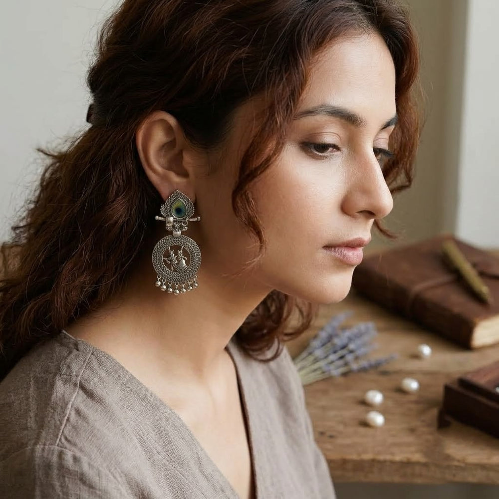 Indian model in rust red outfit wearing Radha-Krishna oxidized silver chandbali earrings, dark bun updo, heritage background