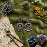 Indian model in grey linen top wearing oxidized silver Radha-Krishna peacock feather chandbali earrings, holding ear, soft light portrait