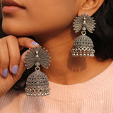 Indian model holding oxidized silver peacock jhumka earring close to face in pink ribbed top, dramatic close-up showing peacock fan detail and granulation dome