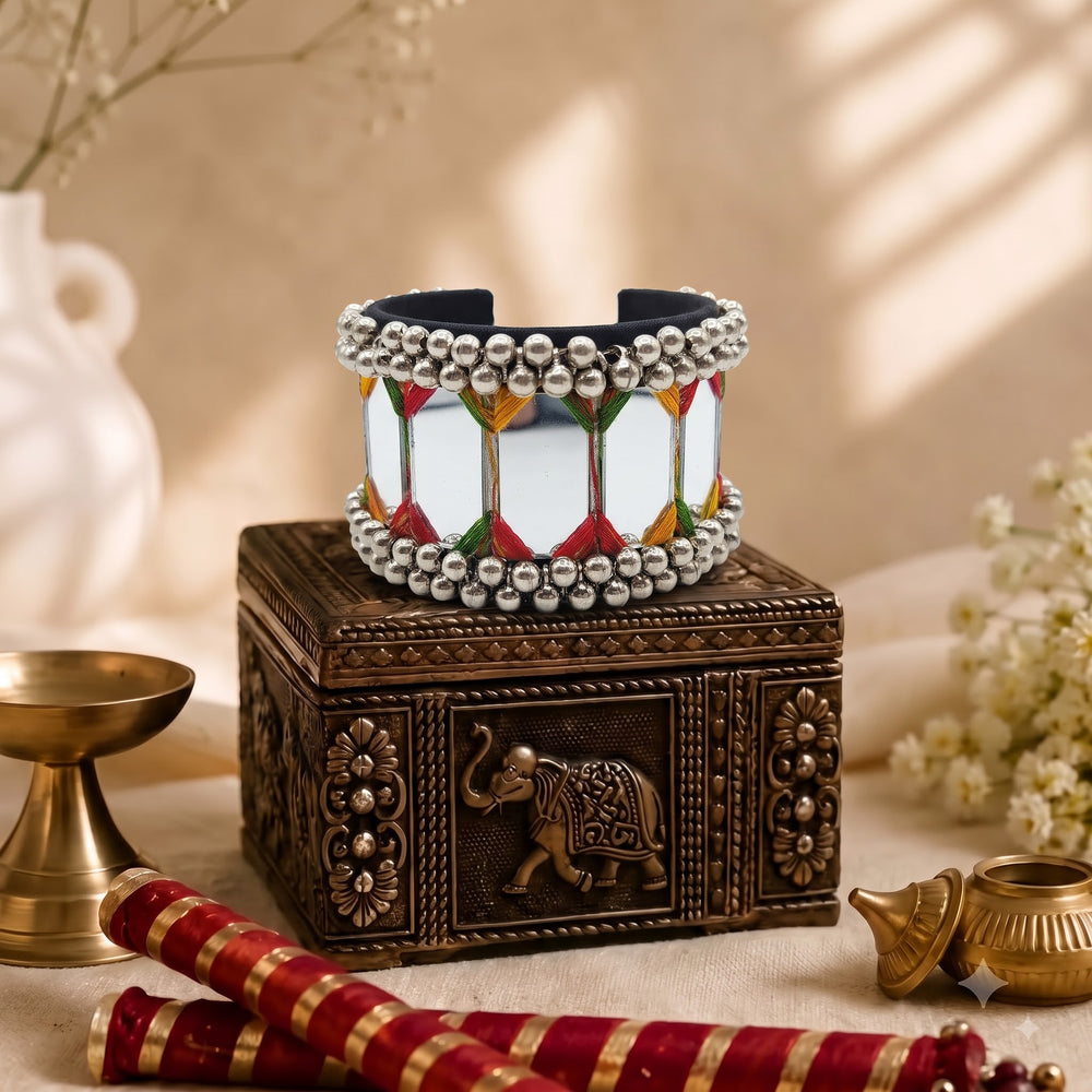 Large mirror tile ghungroo cuff bracelet placed on carved dark wooden jewellery box with brass diya, red and gold bangles and white flowers in warm Diwali-styled heritage flat lay