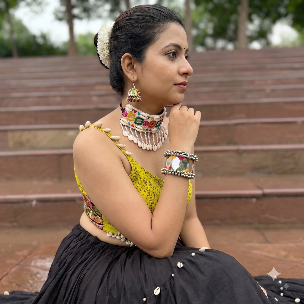 Indian model in traditional Navratri yellow blouse and black skirt wearing large mirror tile ghungroo cuff bracelet, seated editorial pose on outdoor steps with trees in background