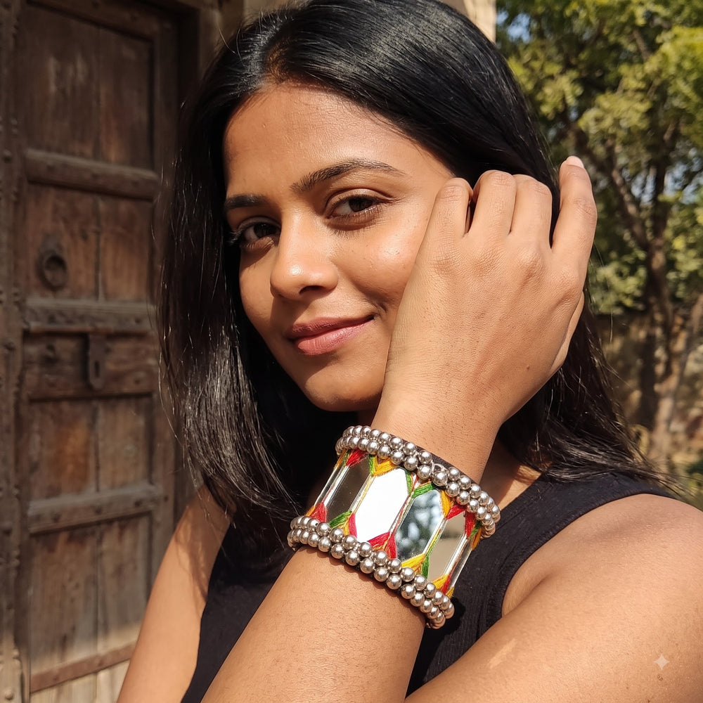 Indian model in yellow Navratri outfit close-up face portrait wearing large mirror tile ghungroo cuff bracelet, hand posed near chin showing bracelet detail with terracotta heritage backdrop