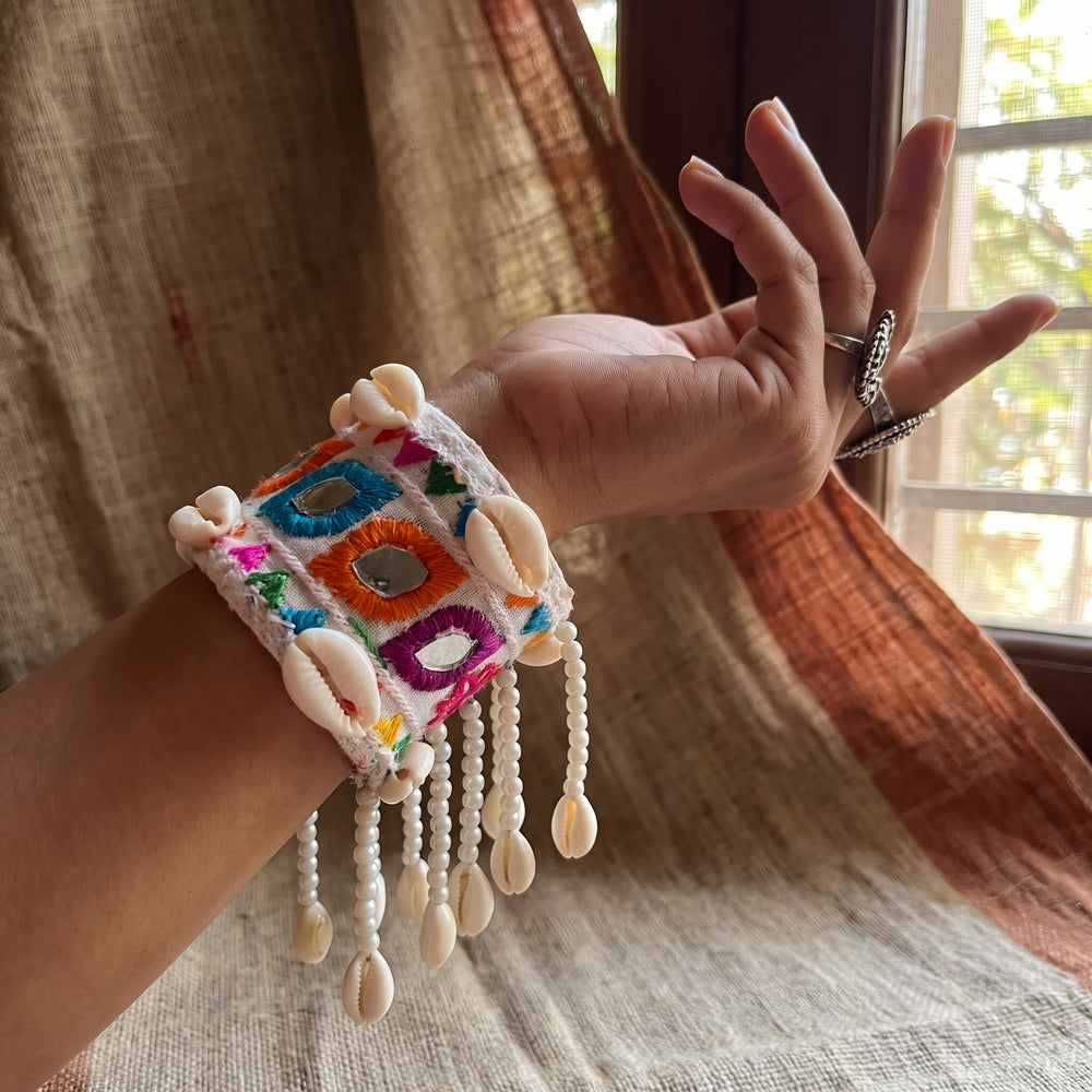 Close-up of hand wearing Kutchi embroidery cowrie cuff bracelet against terracotta wall, showing mirror shisha tiles, multicolour thread diamond motifs and pearl bead cowrie fringe drops in detail