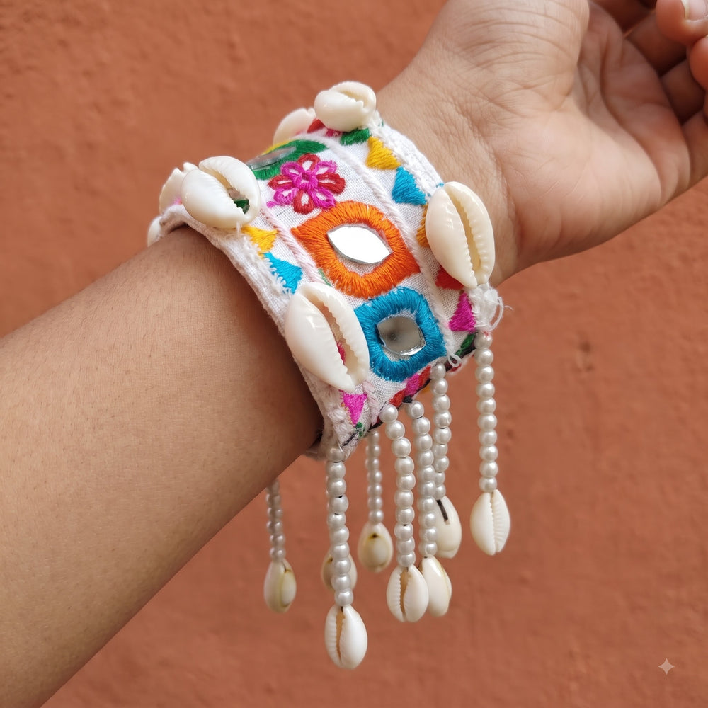 Hand raised near linen drape window wearing Kutchi embroidery mirror work cowrie cuff bracelet, natural light showing full cuff width and swaying pearl bead fringe with cowrie drops