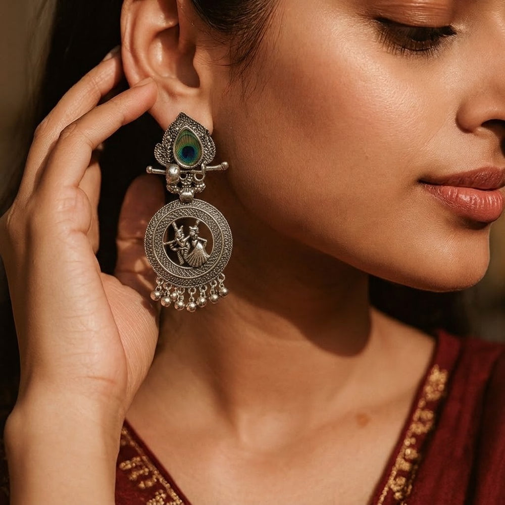Model wearing oxidized Radha-Krishna chandbali earrings with peacock feather, contemplative side profile, lavender and books in background