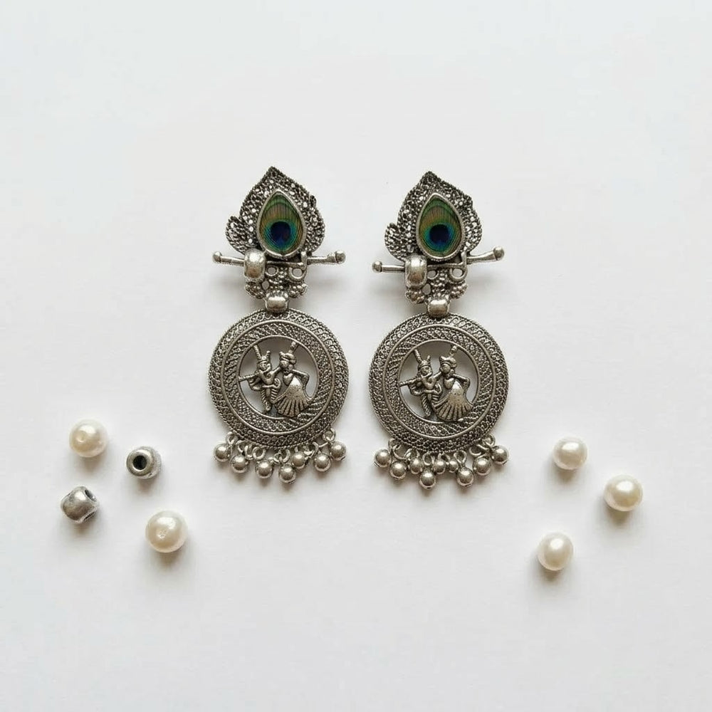 Pair of oxidized silver Radha-Krishna chandbali earrings with peacock feather stud on white background, clean product flat lay with pearls